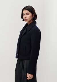 Peacoat scurt de culoare bleumarin, realizat dintr-un material lână texturat, având un guler lat, închidere cu nasturi și buzunare laterale. Modelul poartă pantaloni negri plisați.