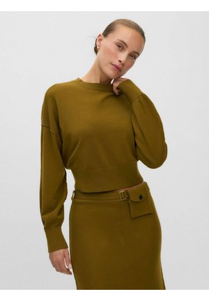 Femme portant un pull en maille vert olive assorti et une jupe avec ceinture et petite pochette avant, posant devant un fond clair uni.