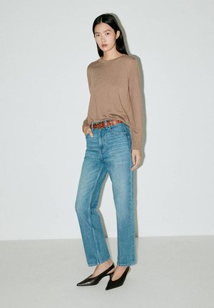 Femme portant un haut marron à manches longues, un jean bleu taille haute avec une ceinture marron, et des talons noirs à bout pointu, debout contre un mur blanc.