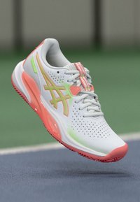 Λευκό αθλητικό παπούτσι με διάτρητη ανώτερη επιφάνεια, παστέλ πράσινες και ροζ λεπτομέρειες, μαξιλαρωτή σόλα και λογότυπο εμφανώς εμφανές στο πλάι.