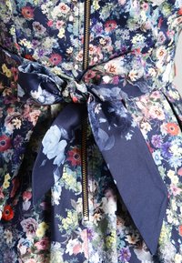 Blumenbekleidungsstück mit einem dunkelblauen Grundton und mehrfarbigen Blumen, einem Reißverschluss auf der Vorderseite und einem stofflichen Gürtel, der mit einer Schleife in der Taille gebunden ist.