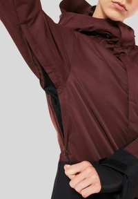 Maroon outdoor jacket with a high collar, zippered sides, mesh lining, and contrasting black accents at the cuffs and hem.
