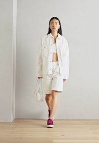 Giacca in denim bianco e pantaloni shorts abbinati a un top corto, insieme a una borsa bianca e sneakers viola. Texture liscia, vestibilità rilassata, design minimale.
