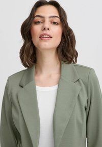 Ljusgrön blazer med en skräddarsydd design, förädlad med en reverskrage och mjuk textur, bärs över en vit ribbad topp.
