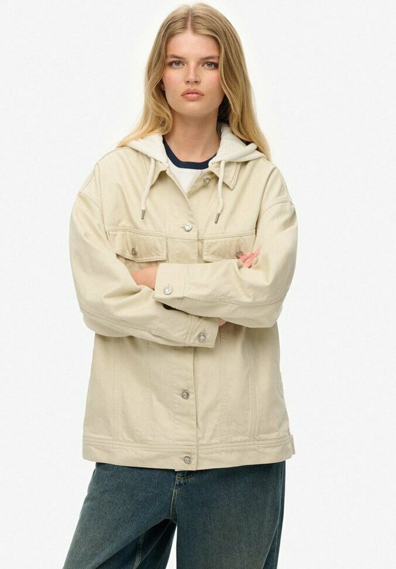 Ung kvinde med langt blond hår iført en oversized beige jakke med hætte og blå jeans, arme krydset, neutral ansigtsudtryk.