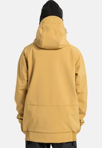 Persoon in een mosterdgeel hoodiejack en een zwarte gebreide beanie, van achteren zichtbaar tegen een effen lichte achtergrond.