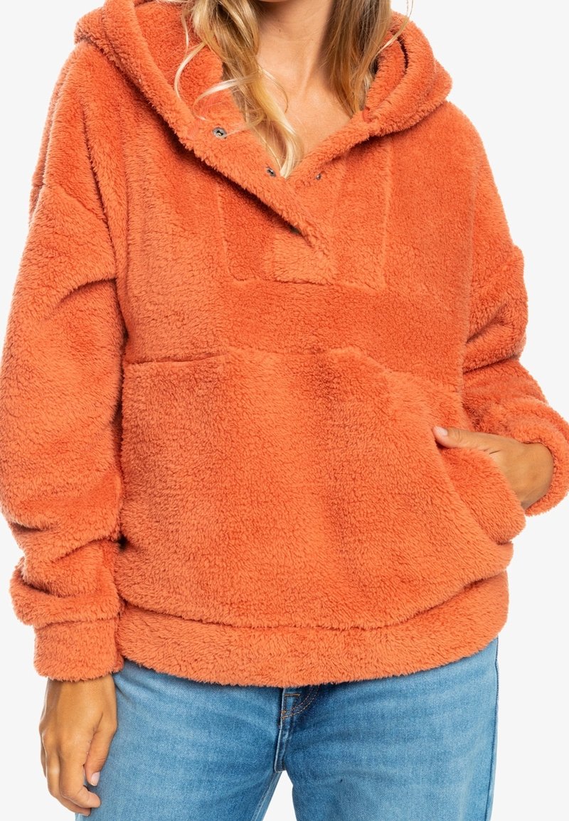 Personne portant un pull à capuche orange pelucheux avec poche avant et jean bleu clair, main partiellement dans la poche, visible des épaules aux hanches.