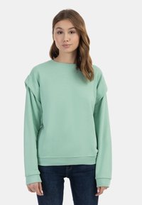Felpa oversize verde menta con maniche lunghe e scollatura rotonda, realizzata in tessuto morbido con polsini e orlo a coste. Indossata con jeans scuri.