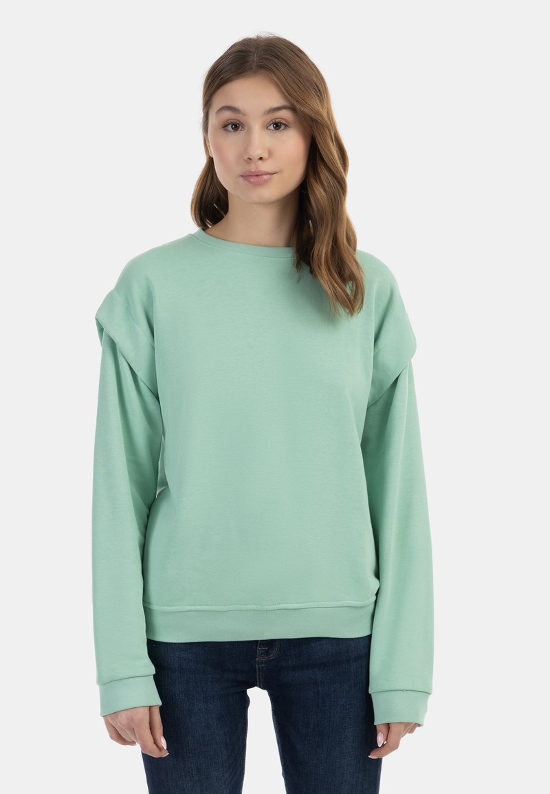 Felpa oversize verde menta con maniche lunghe e scollatura rotonda, realizzata in tessuto morbido con polsini e orlo a coste. Indossata con jeans scuri.