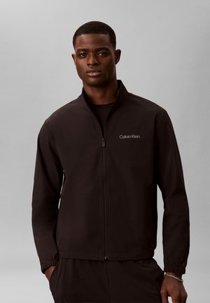 Ung mand iført en sort Calvin Klein jakke med lynlås og hænderne i lommerne mod en ensfarvet grå baggrund.