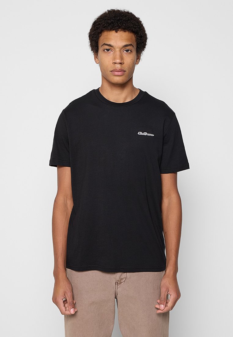 Jonge man die staat, gekleed in een eenvoudig zwart T-shirt met korte mouwen met kleine witte tekst op de borst en lichtbruine broek.