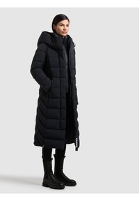 Lunga giacca a vento nera con cappuccio, caratterizzata da un motivo trapuntato, chiusura con zip e tasche laterali. Indossata con pantaloni neri e stivali alla caviglia.