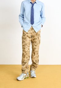 Beżowe spodnie typu cargo w kamuflażu z kieszeniami, w połączeniu z jasnoniebieską koszulą z kołnierzykiem i granatowym krawatem w paski, do tego szaro-zielone buty sportowe.