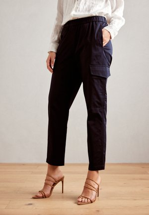Marineblaue Hose mit elastischem Bund, Seitentaschen, tailliertem Schnitt und glatter Textur. Kombiniert mit beigen, strappy High-Heels.