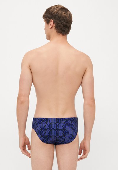 Männliches Model von hinten gesehen, das blaue Schwimmbrieftaschen mit einem kräftigen, wiederholten "DSQUARED2"-Logomuster auf einem schlichten weißen Hintergrund trägt.