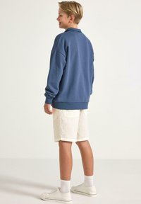 Blå sweatshirt med en avslappnad passform, ribbade muddar och nederkant, bärs över vita shorts. Vita sneakers kompletterar outfiten. Bakgrunden är neutral.