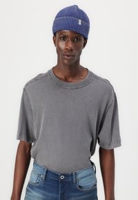 Grå t-shirt med rund hals i mjukt material, med korta ärmar och avslappnad passform. Bärs med blå jeans och en ribbad blå mössa.