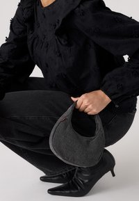 Grå handväska med en böjd form, bärs av en person i svart texturerad blus och mörkgrå jeans, som lyfter fram spetsiga svarta klackar.