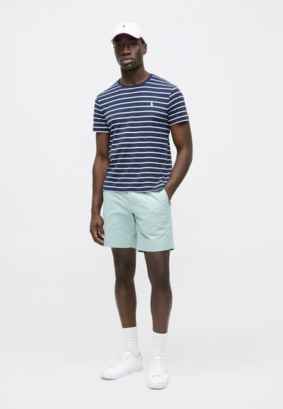 Μπλε ναυτικό ριγέ μπλουζάκι με κοντά μανίκια, σορτς σε απόχρωση μέντας, λευκά αθλητικά παπούτσια και ροζ καπέλο με λογότυπο. Καθημερινό καλοκαιρινό ντύσιμο σε έκθεση.