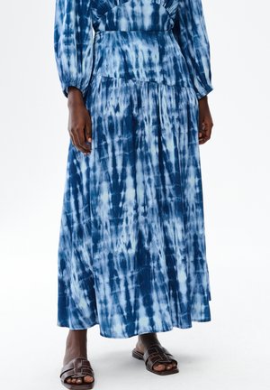 Niebiesko-biała sukienka maxi tie-dye noszona z brązowymi sandałami typu slip-on z odkrytymi palcami, pokazana od połowy klatki piersiowej do stóp na osobie o ciemnej karnacji.