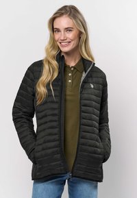 Svart vadderad jacka med vertikal quiltad struktur, dragkedja framtill och lätt isolering. Buren över en grön pikétröja med upprättstående krage.