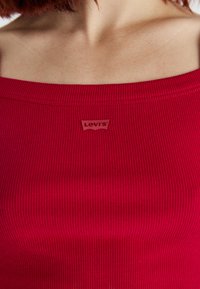 Top de tanque vermelho com textura canelada e decote largo, apresentando uma pequena etiqueta em relevo do logo Levi's perto da área do peito. Textura suave.