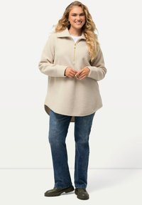 Pullover beige in fleece con zip parziale, colletto alto e orlo curvo. Abbinato a jeans in denim blu e scarpe slip-on nere.
