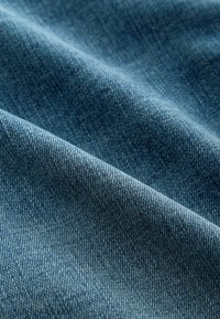 Gros plan d'un tissu denim bleu avec une texture de tissage diagonal visible et des plis doux créant ombres et lumières.