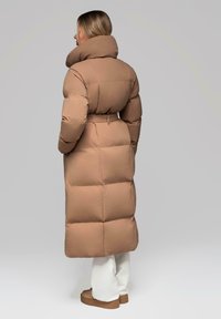 Žena v dlouhém hnědém prošívaném kabátu s páskem a vysokým límcem, bílých kalhotách a hnědých botách, stojící proti šedému pozadí, odvrácená.