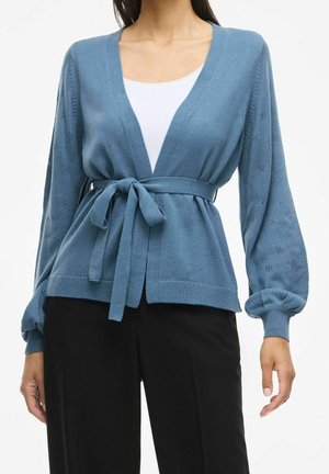 Cardigan bleu en maille avec détails ajourés sur les manches ballon, ceinture nouée à la taille, porté sur un haut blanc et un pantalon noir.