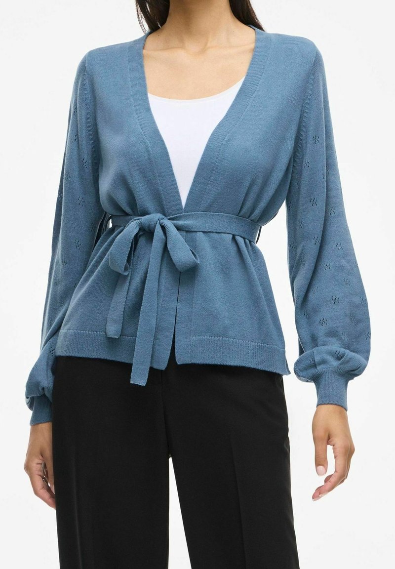 Cardigan bleu en maille avec détails ajourés sur les manches ballon, ceinture nouée à la taille, porté sur un haut blanc et un pantalon noir.