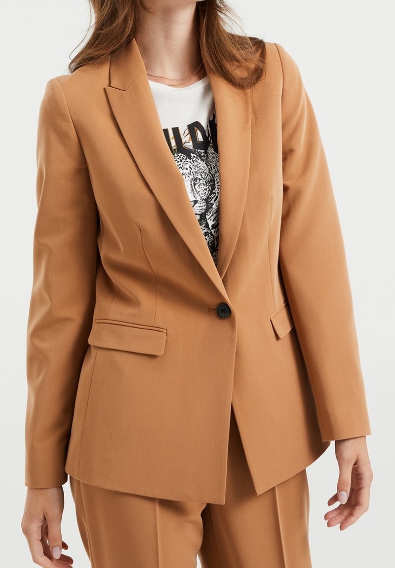 Femme portant un blazer ajusté couleur camel sur un t-shirt blanc graphique avec un imprimé de visage de léopard, assorti d'un pantalon coordonné.