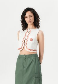 Witte cropped top met oranje geweven bies en een zon-embroidery, met een knoopsluiting en een enkele borstzak. Gepaard met groene cargorok.