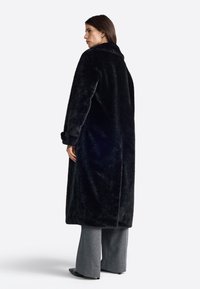 Zwarte neppelsveten lange jas met een kraag, zijzakken en brede mouwen. Gladde textuur en een ontwerp met volle lengte, gedragen over grijze wijde broek.