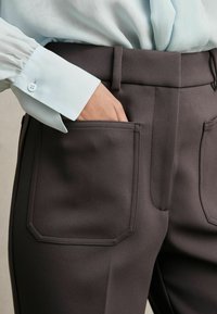 Pantaloni di colore carbone con una vestibilità su misura, caratterizzati da due grandi tasche e un tessuto leggermente strutturato. Abbinati a una camicia azzurro chiaro con polsini a bottoni.