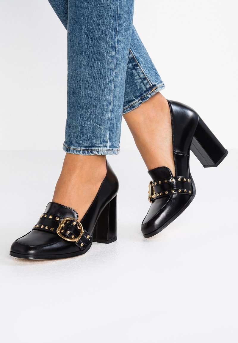 Personne portant un jean bleu et des chaussures à talons noirs avec boucle dorée et détails cloutés sur fond blanc.