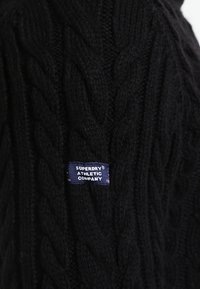 Pull noir en maille torsadée avec un motif texturé, arborant une étiquette bleue portant l'inscription "SUPERDRY ATHLETIC COMPANY" cousue sur le côté.