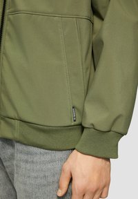 Olivegrüne Jacke mit glatter Textur, gerippten Bündchen und Seitentaschen. Besitzt ein Markenlabel, das in die Naht eingestickt ist.