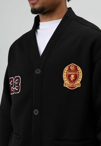 Zwarte cardigan met een V-hals, twee knopen, steekzakken aan de voorkant en borduurdetails in bordeauxrood en goud, waaronder het nummer 23 en een logo.