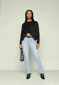 Noor naine punutud juustega, kellel on seljas must lühike tops, avatud must bleiser, helesinised teksad ja mustad saapad, hoiab musta käekotti külje juures.