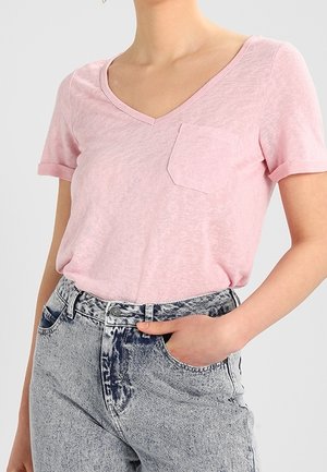 Woman wearing a light pink V-neck T-shirt with a small chest pocket, tucked into high-waisted acid-wash blue jeans with a hand in the pocket.