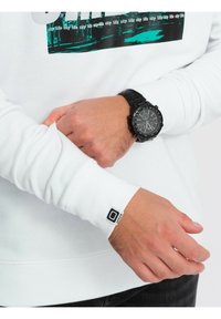 Biały sweter z nadrukiem graficznym przedstawiającym turkusowy i czarny wzór. Czarny zegarek na nadgarstku. Długie rękawy z mankietami z detalem marki.