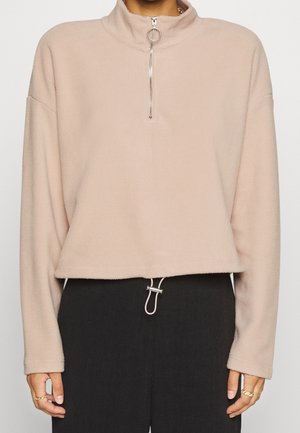 Pull à polaire beige avec une demi-fermeture éclair et un tirant en anneau, associé à un pantalon noir avec une boucle argentée à la taille.