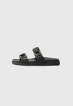Black leather slide sandals with two wide straps featuring gold circular buckles, a flat textured sole, and a minimalist design.