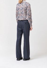 Femme portant un pantalon large bleu marine à rayures beiges verticales, associé à une blouse à manches longues à motifs et des chaussures noires épaisses, se tenant de dos.