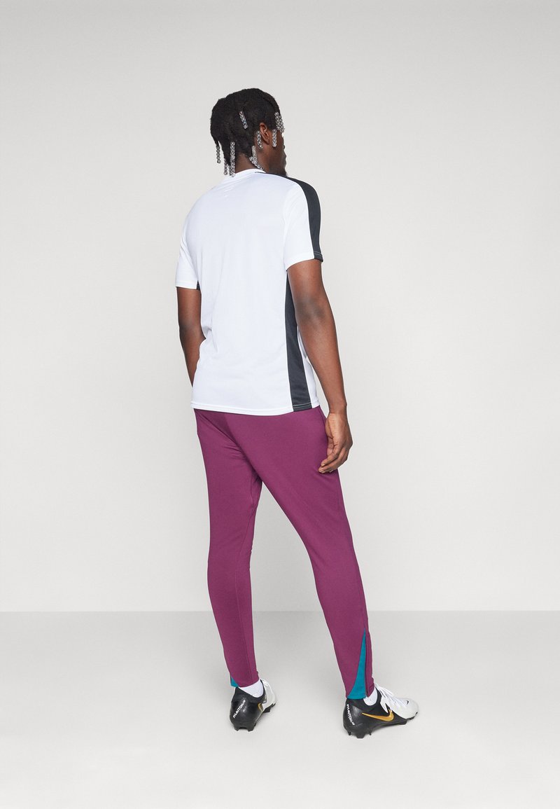 T-shirt blanc de sport avec des panneaux latéraux noirs, associé à un pantalon violet ajusté avec des accents turquoise au niveau de l'ourlet. Les chaussures de sport sont noires et blanches.
