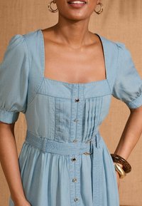 Light blue denim dress features a square neckline, puffed sleeves, front pleats, button closures, and a matching belt with a textured finish.