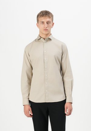 Jeune homme se tenant face à l'avant, portant une chemise beige à boutons et un pantalon noir, sur un fond blanc uni.