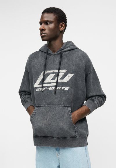 Fiatal férfi, aki egy kifakult fekete kapucnis pulóvert visel fehér „OFF-WHITE” feliratú grafikával és világoskék farmert, kezét a pulóver zsebében tartja, balra néz.