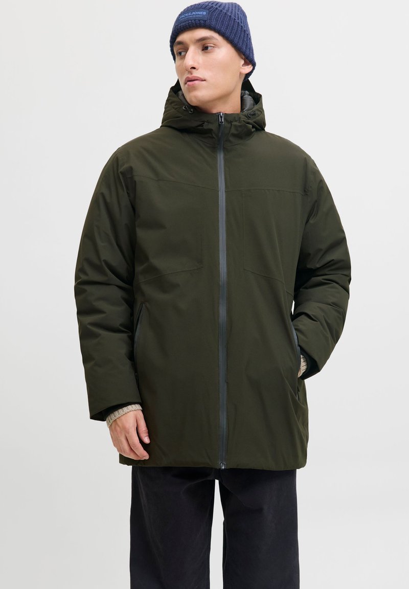 Mees, kes kannab tumerohelist kapuutsiga talvejope ja tumesinist mütsi, seisab ühe käega taskus ja vaatab küljele.